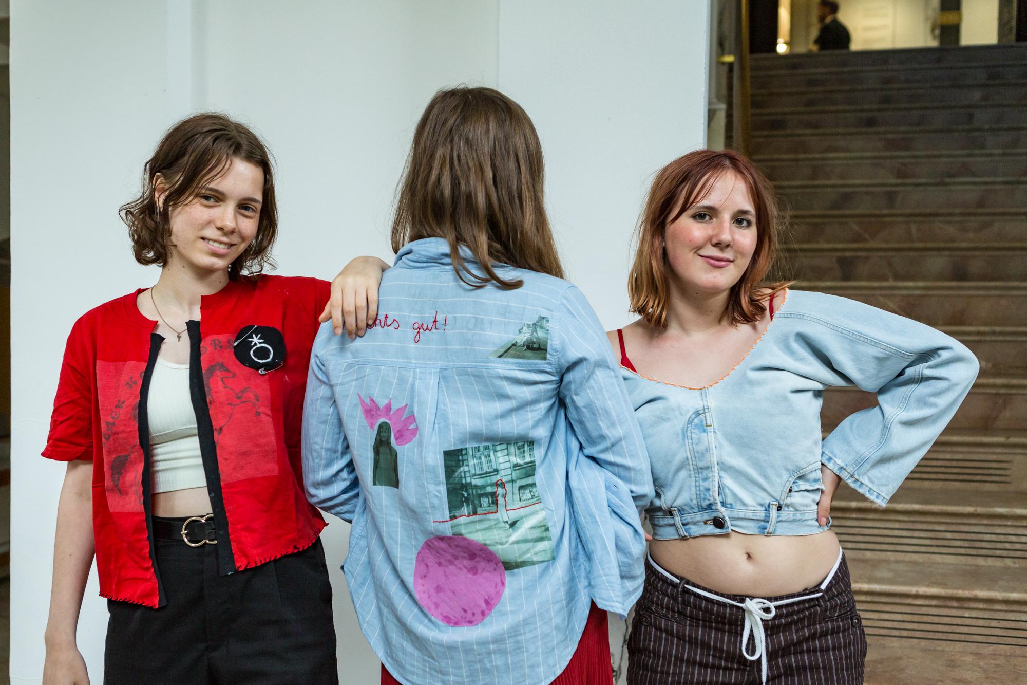 Three young, female-read people stand in a row and present their embroidered clothes. The person in the middle turns her back to the camera.
