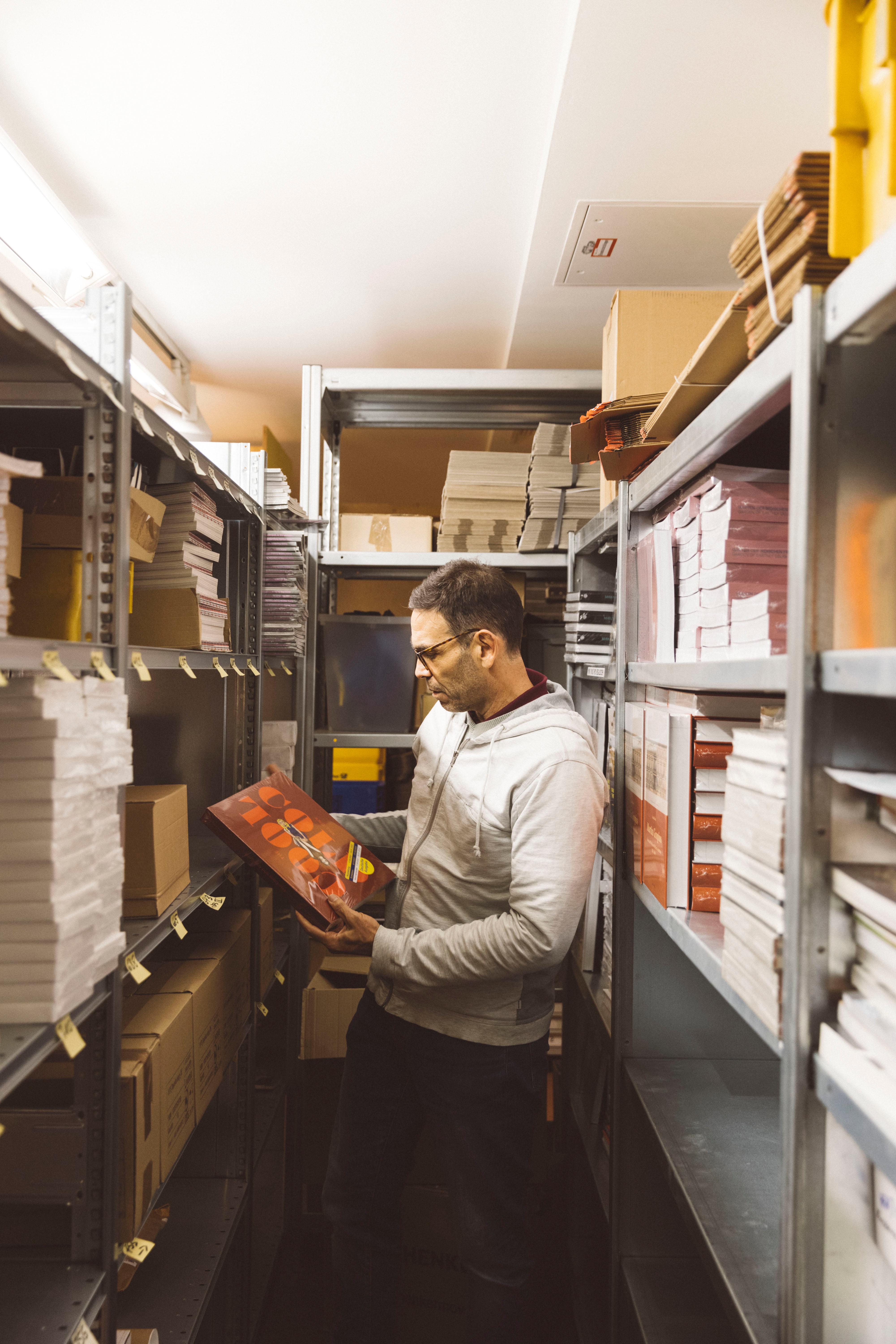 A person stands between shelves with magazines and books and holds an exhibition catalog in his hands.