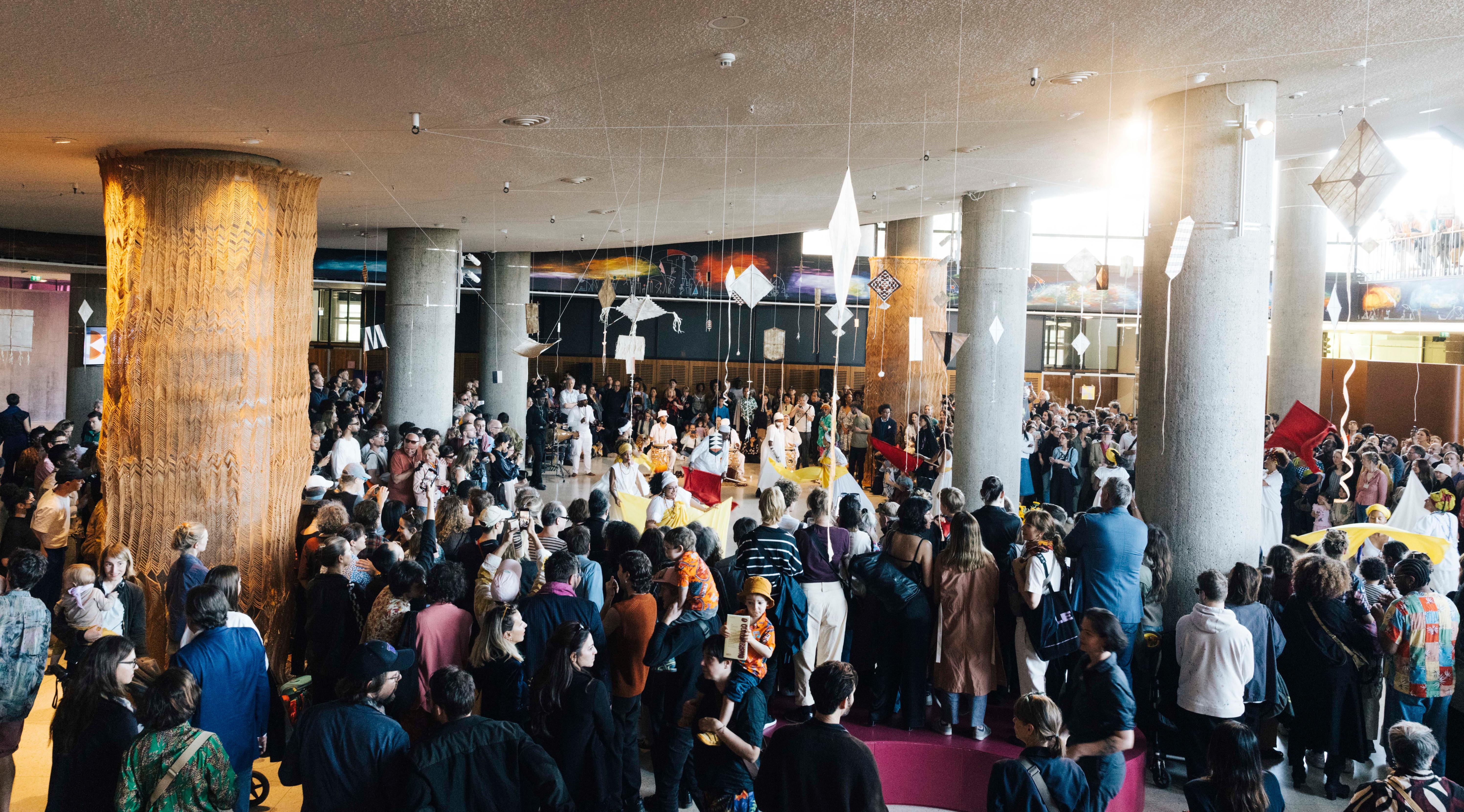 Many people stand in a room with pillars. In the middle there is a performance with dancers and musicians.