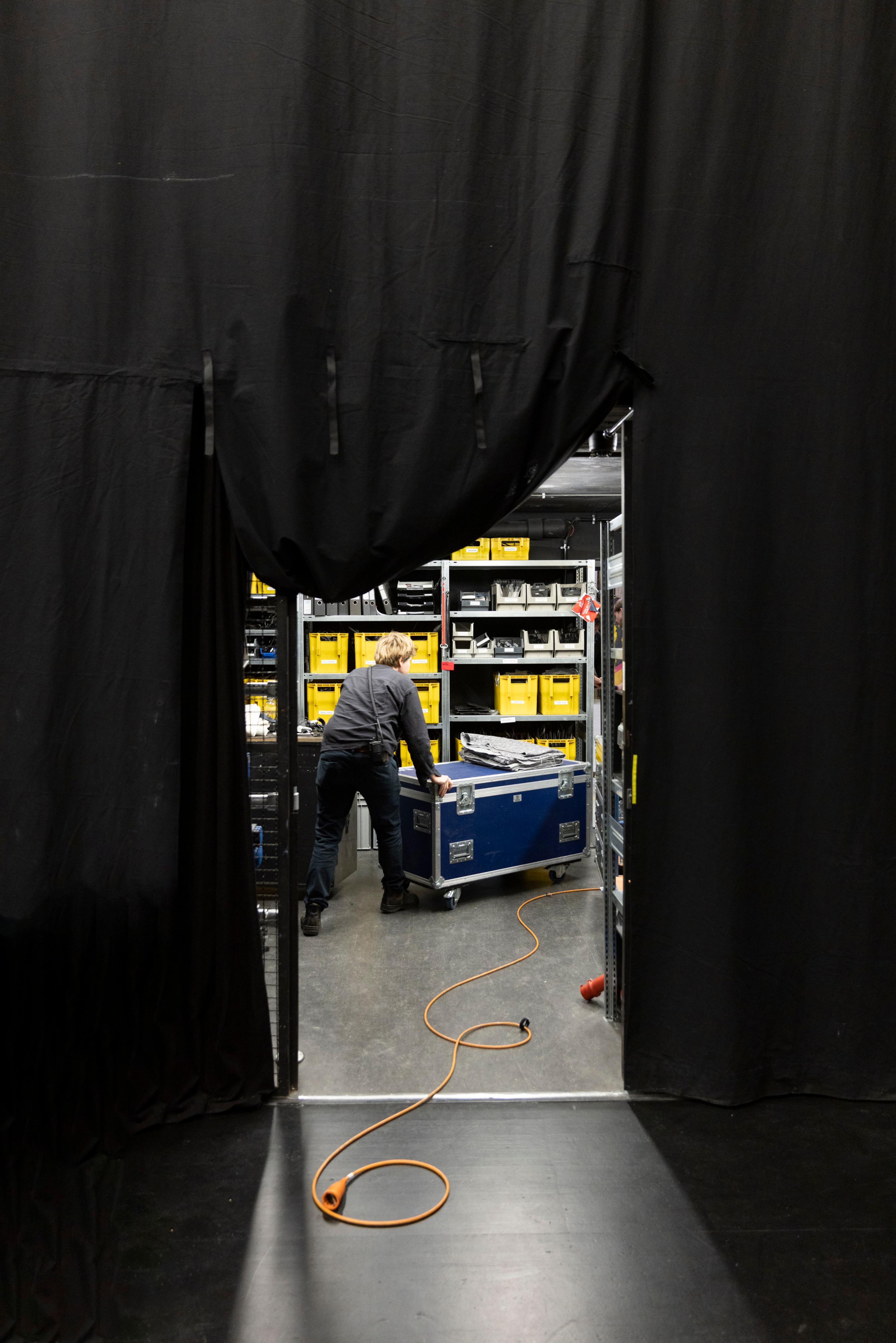 A person pushes a container in a small room. The entrance to the room is flanked by a black curtain.