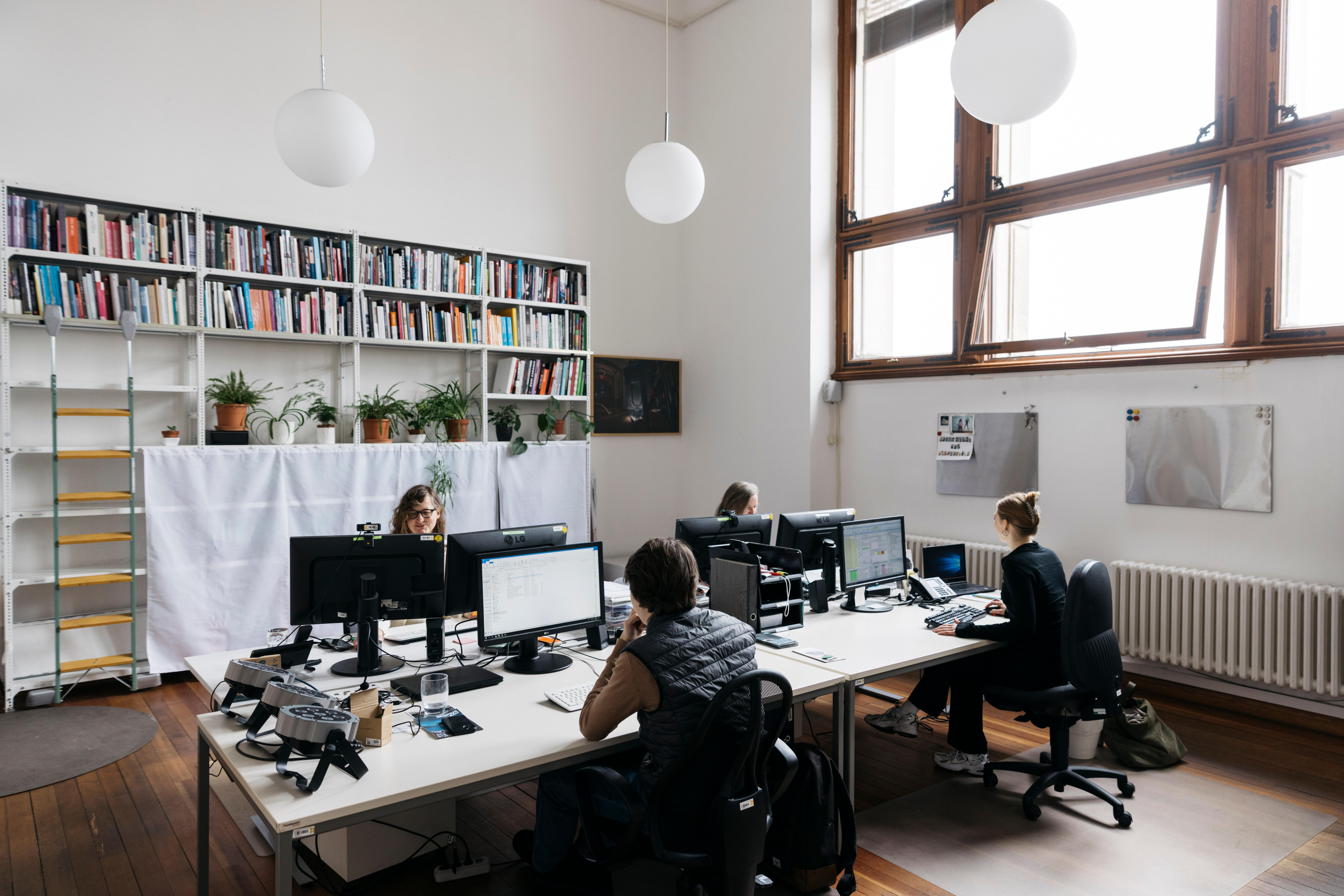 Employees are sitting at their desks in the office. The office is bright, there is a large bookshelf on the back wall.