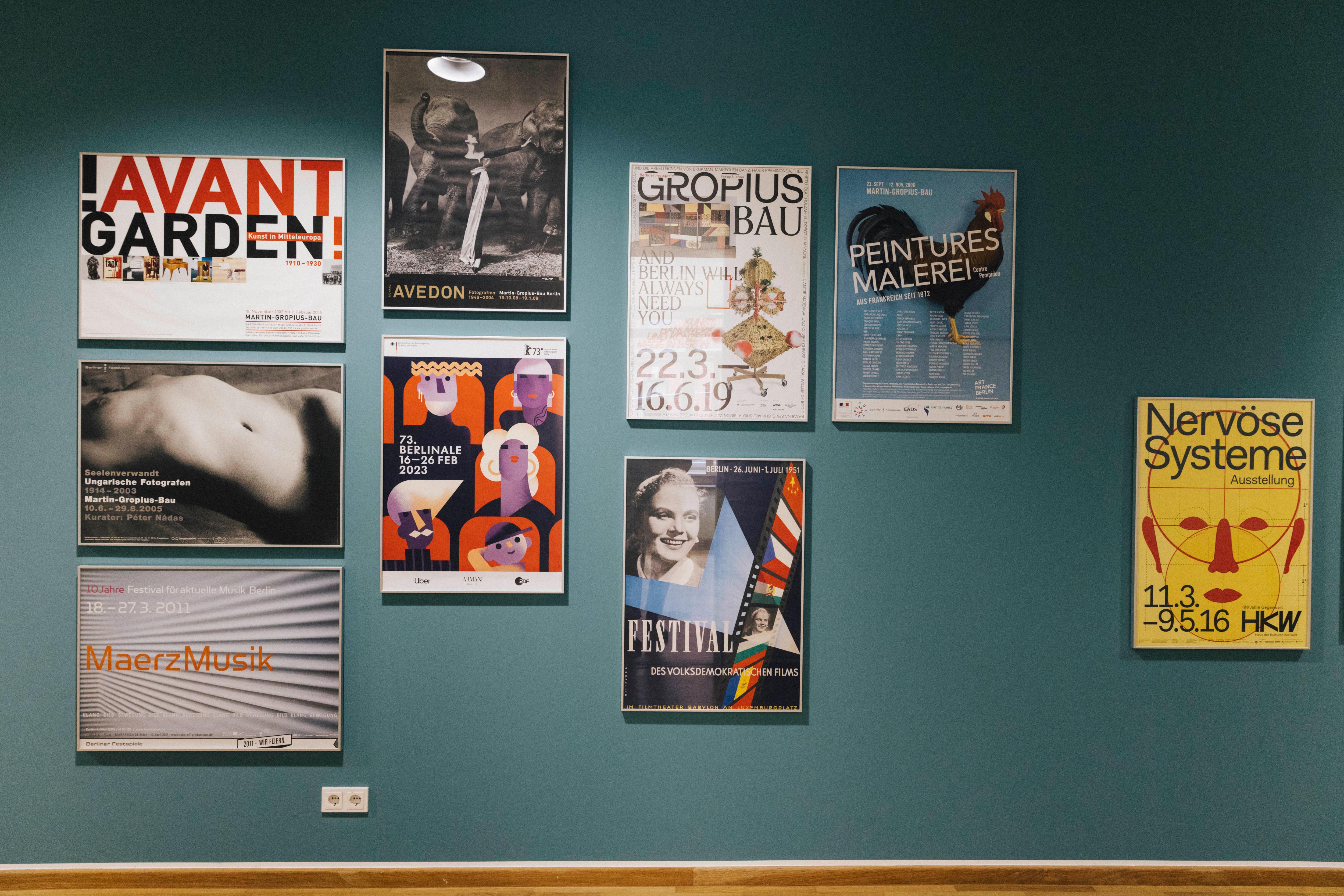 Framed exhibition and event posters hang on a petrol-coloured wall.