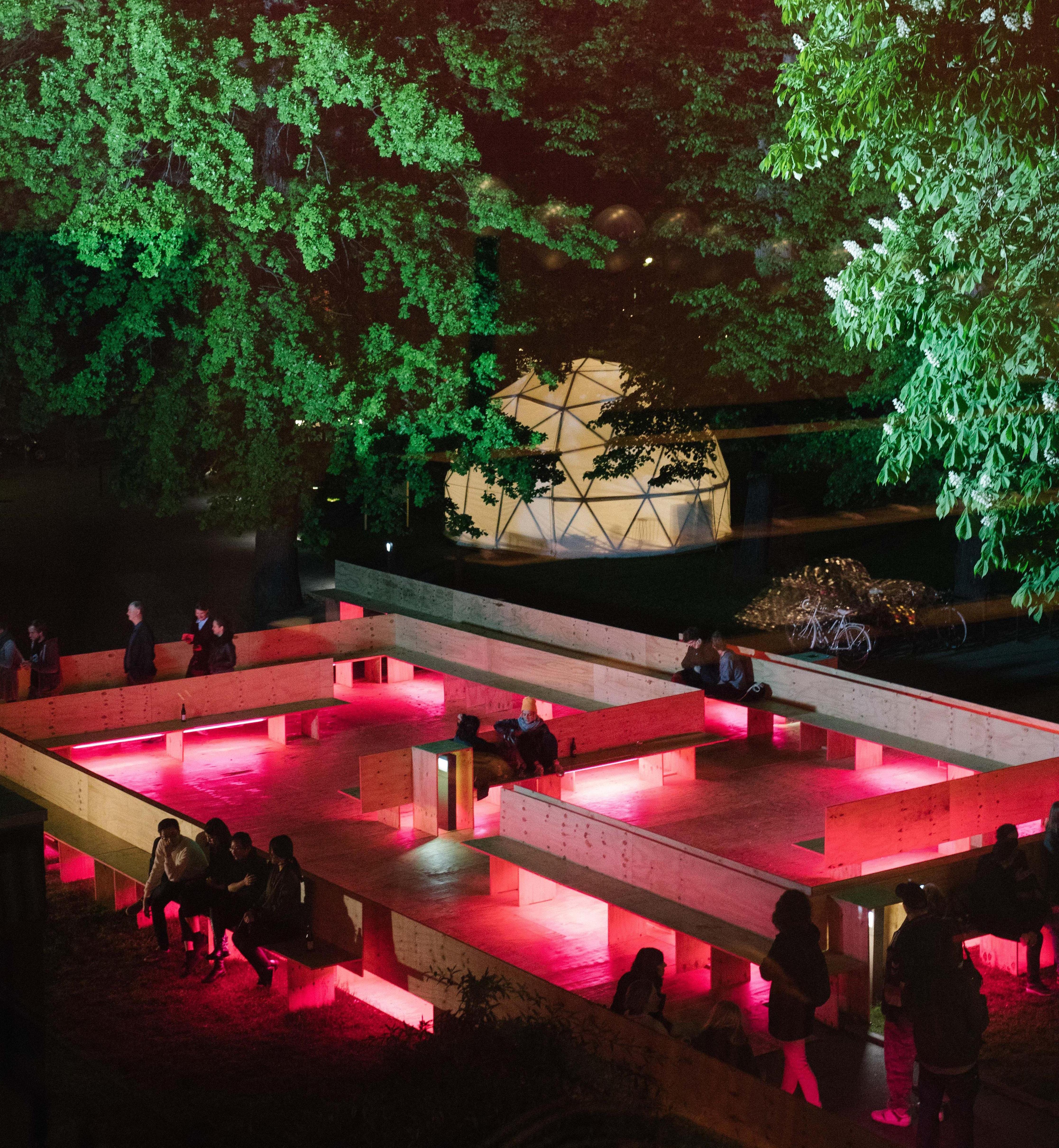 Wooden benches on the forecourt of the Haus der Berliner Festspiele are arranged like a maze and illuminated by pink light at night.