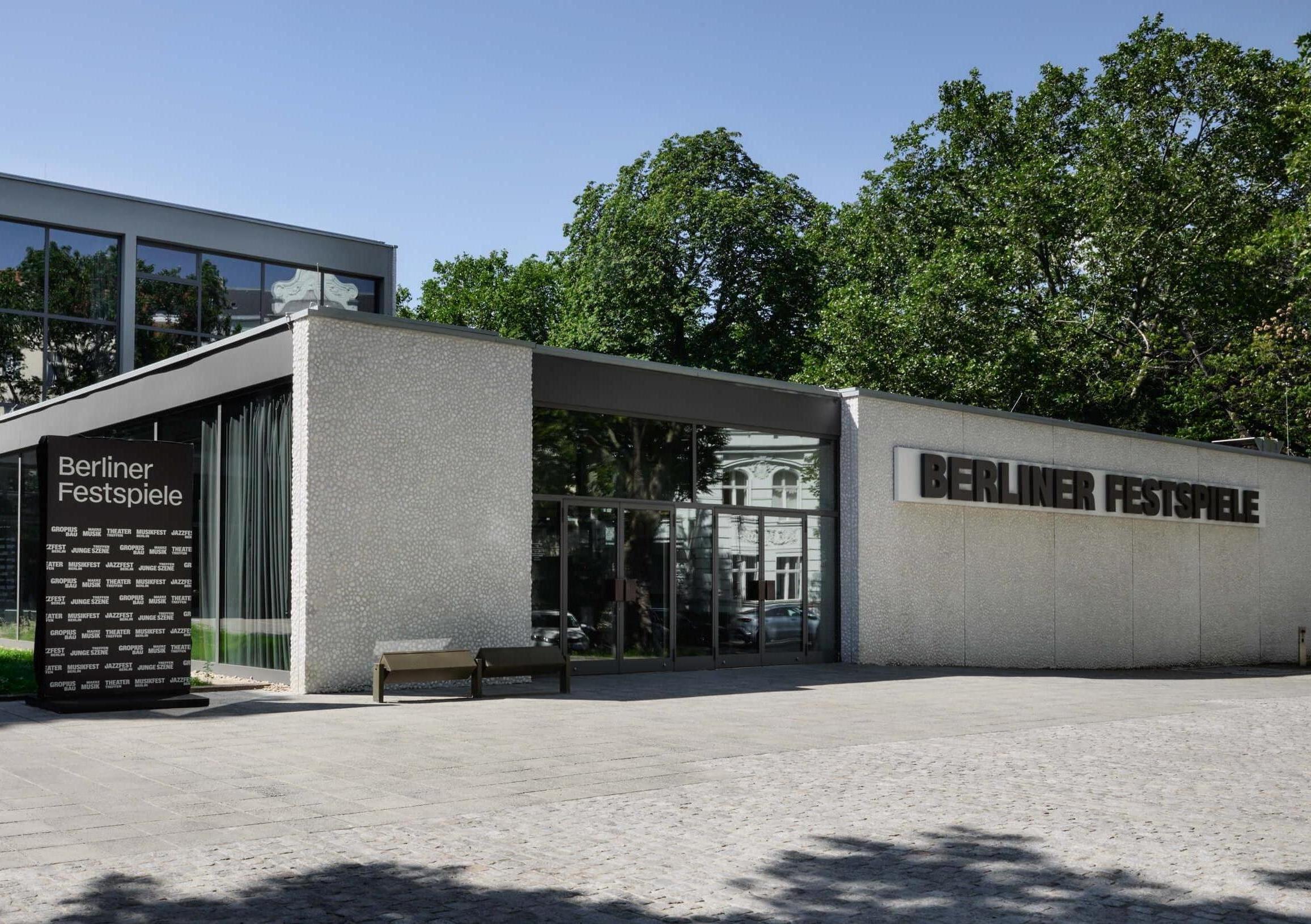 Frontal view of the Haus der Berliner Festspiele. The white building of the box office hall with the lettering "Berliner Festspiele" and the large glass façade of the front building can be seen.