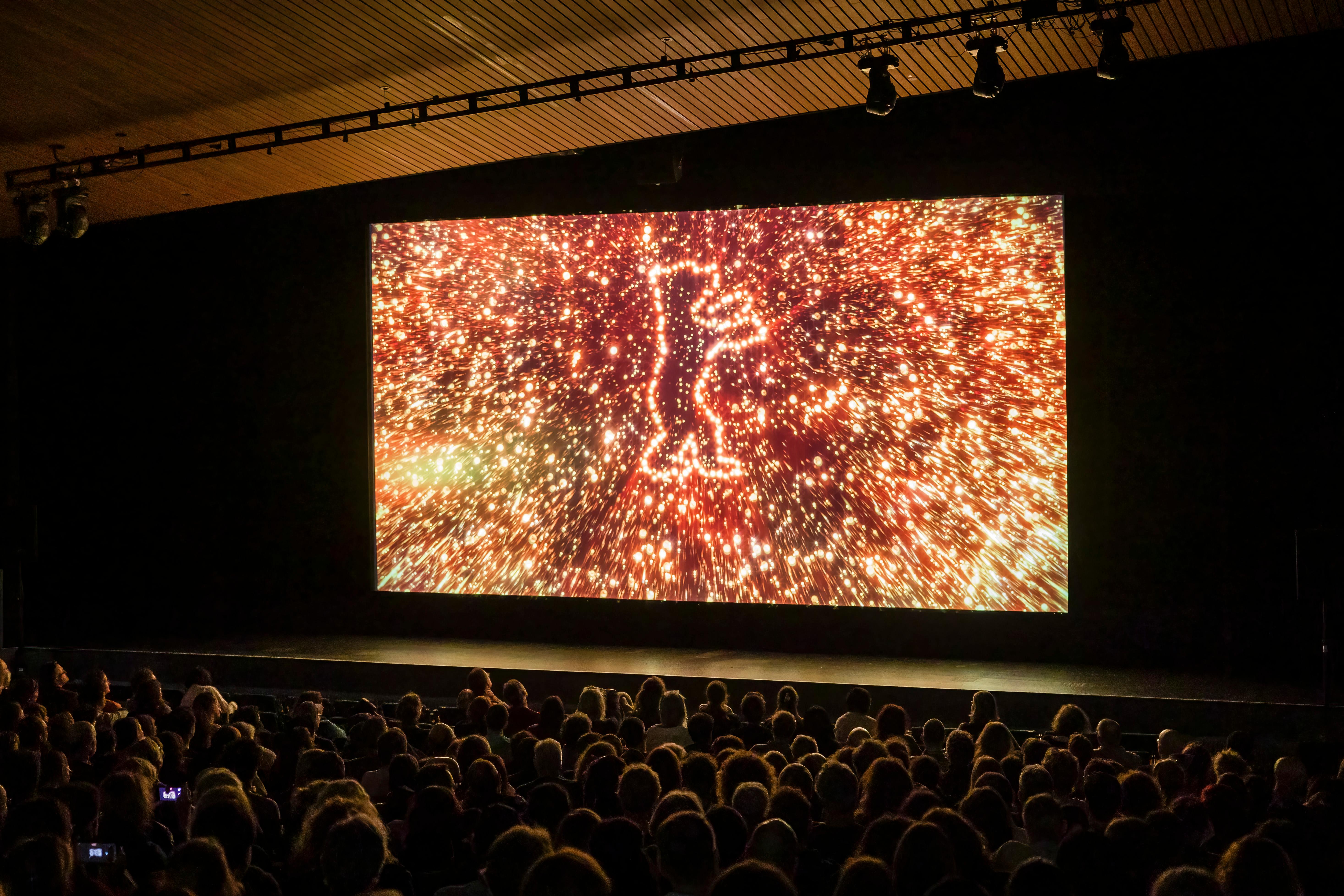 The cinema hall of the Akademie der Künste with audience. The trailer of the Berlinale can be seen on the screen. The trailer shot shows the Berlinale Bear in the glowing fireworks.