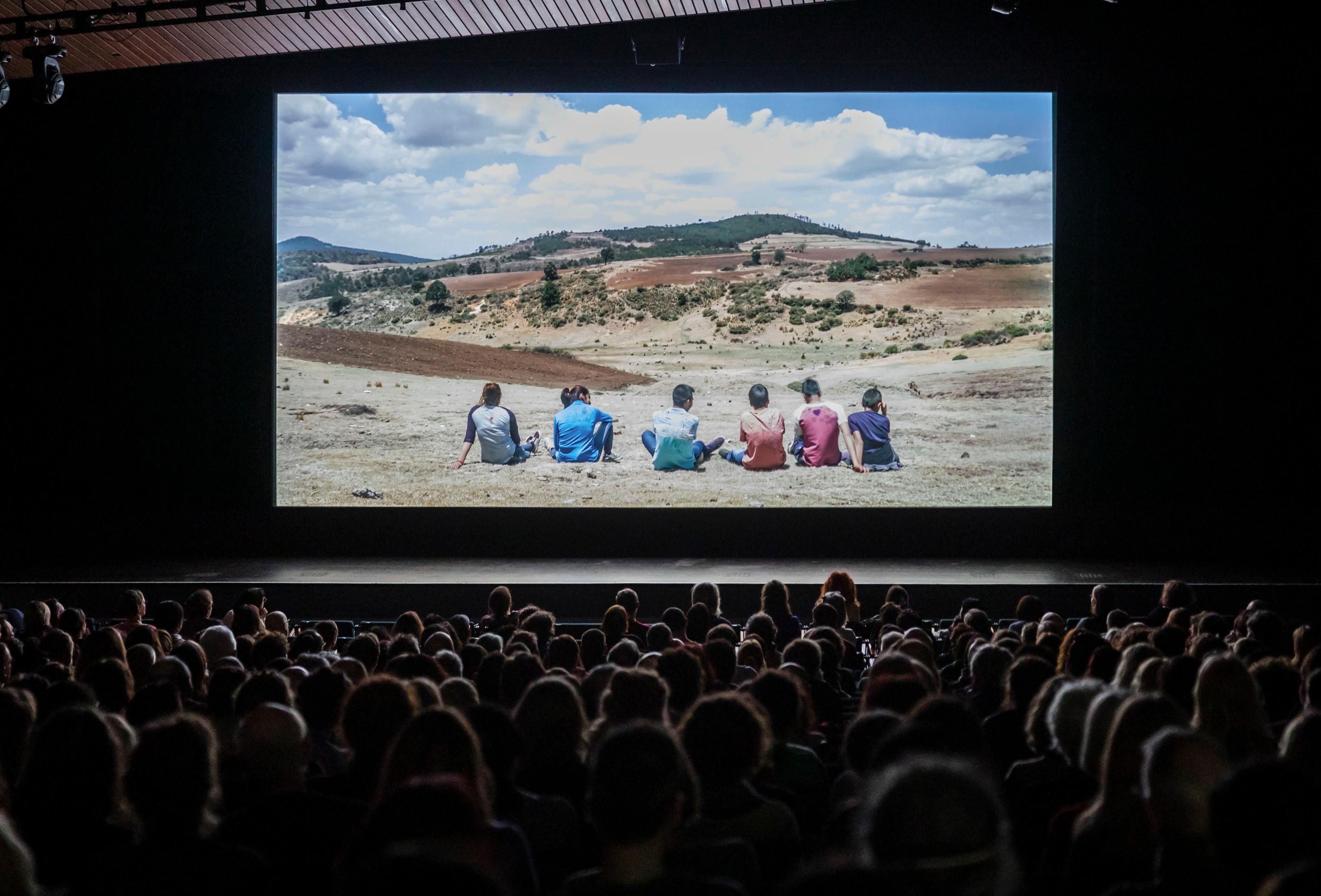 Der Kinosaal der Akademie der Künste mit Publikum. Auf der Leinwand ist der Film El eco zu sehen. Die Szene zeigt eine Gruppe Jugendlicher, die auf dem Boden in einer Prärie sitzen.
