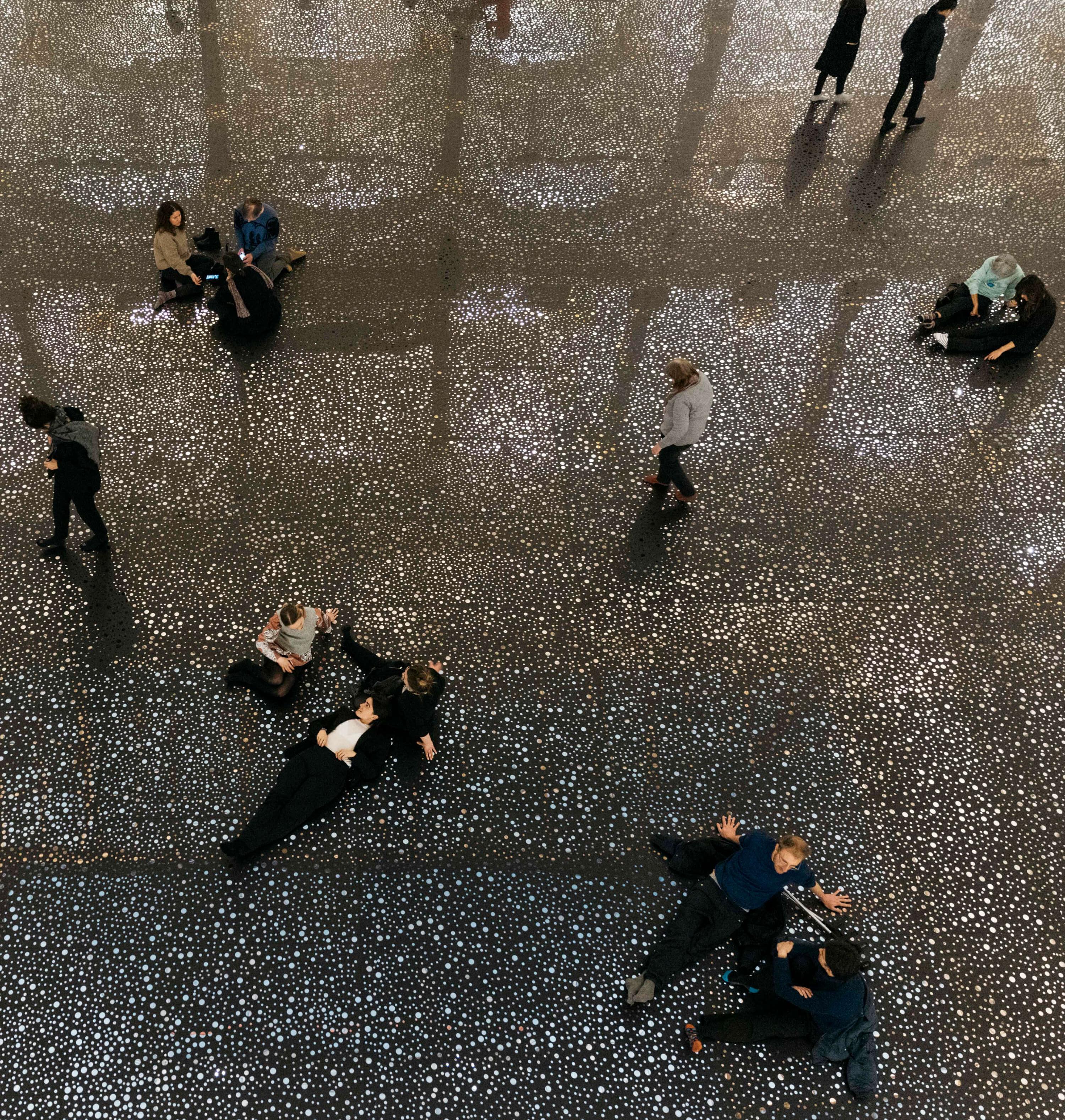 The atrium of the Gropius Bau. The floor is covered with a reflective foil as part of the exhibition "RAINBOW SERPENT (VERSION)" by Daniel Boyd. Visitors sit and lie on the installation.