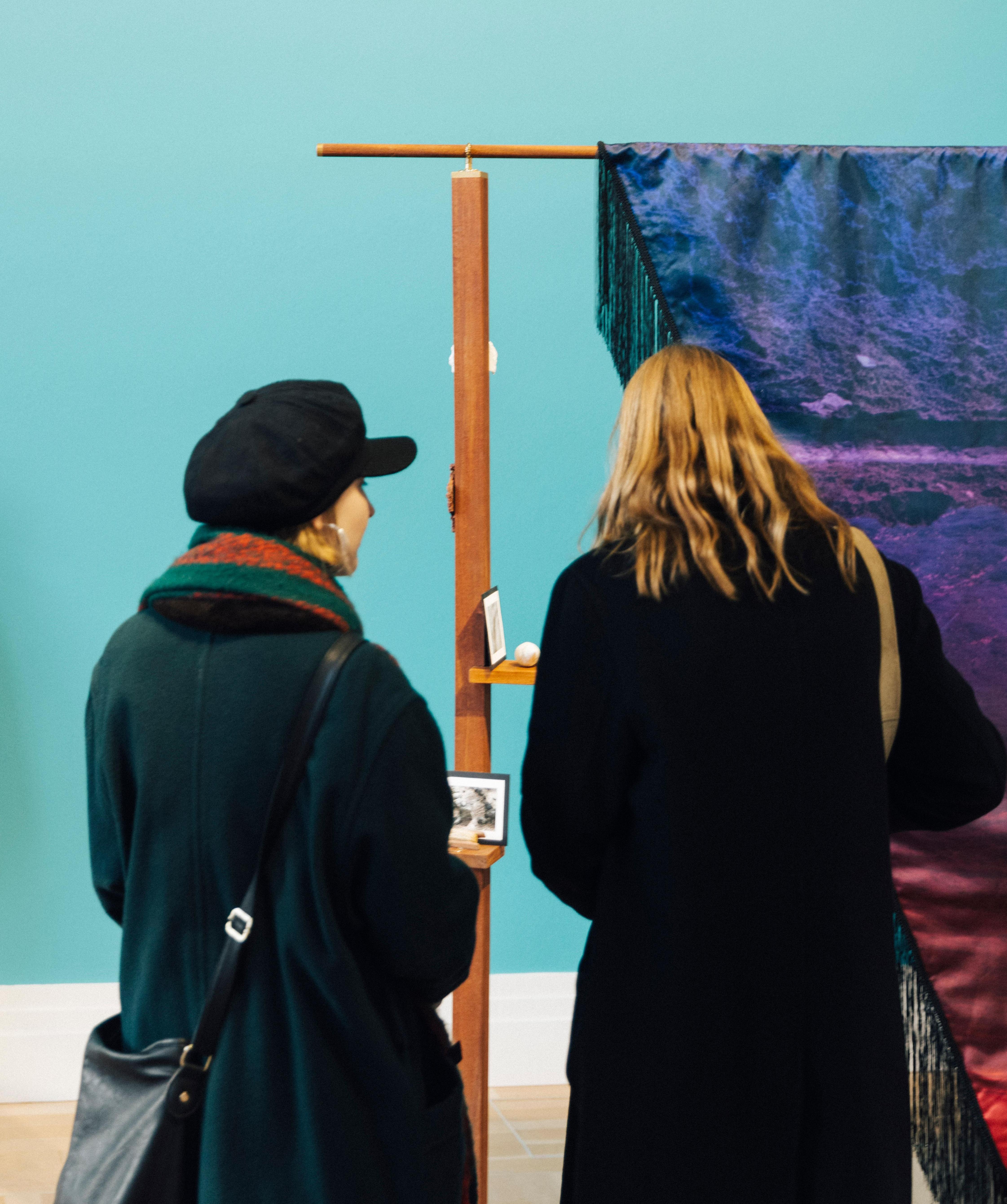 Two people are looking at a work by the artist Shiraz Bayjoo. The sculptural work consists of textile flags. The wall in the background is turquoise blue.