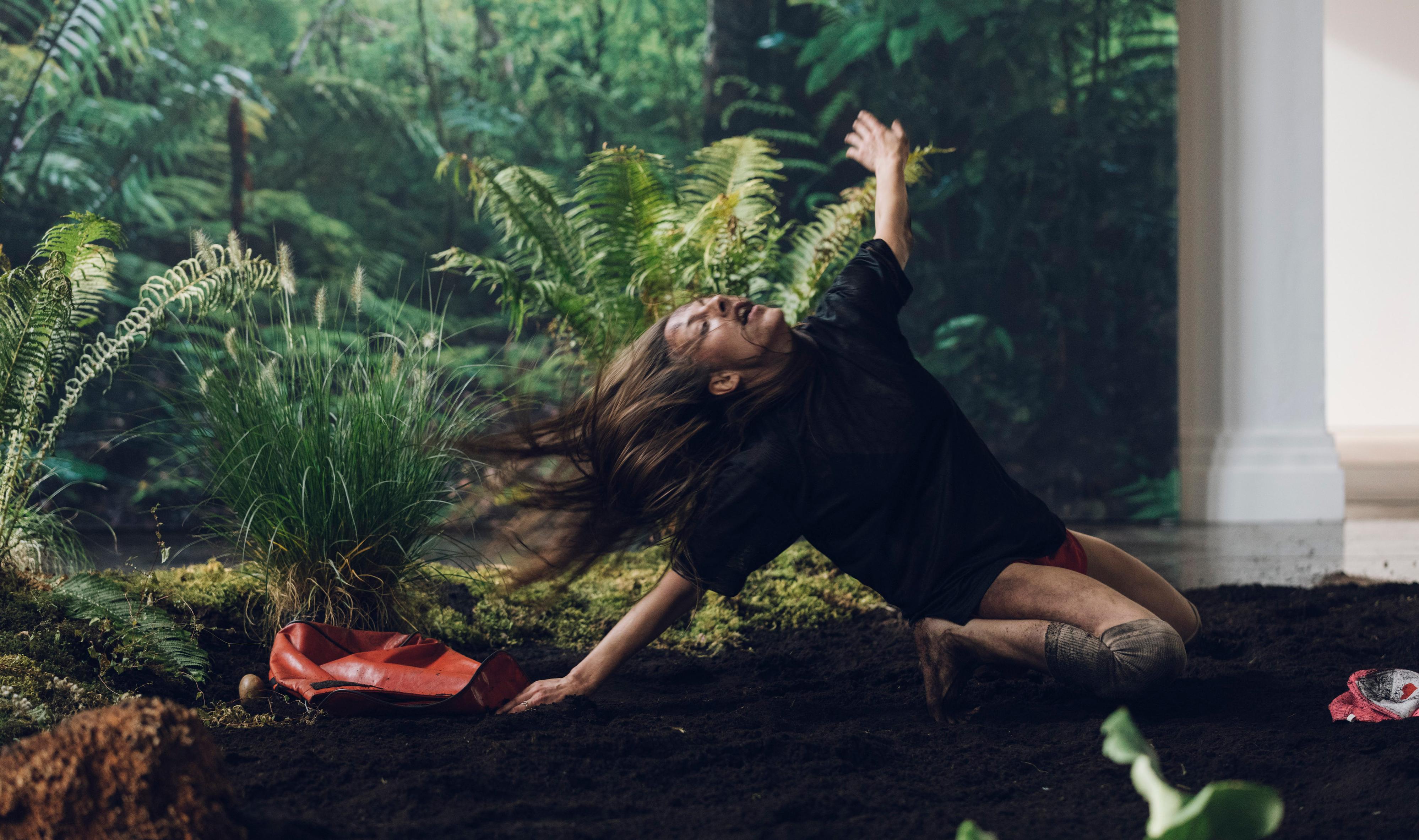 Eine weiblich gelesene Person kniet in einem Raum auf dem Boden, der mit Erde bedeckt ist. Im Hintergrund sind Pflanzen und eine Tapete mit Waldmotiv.