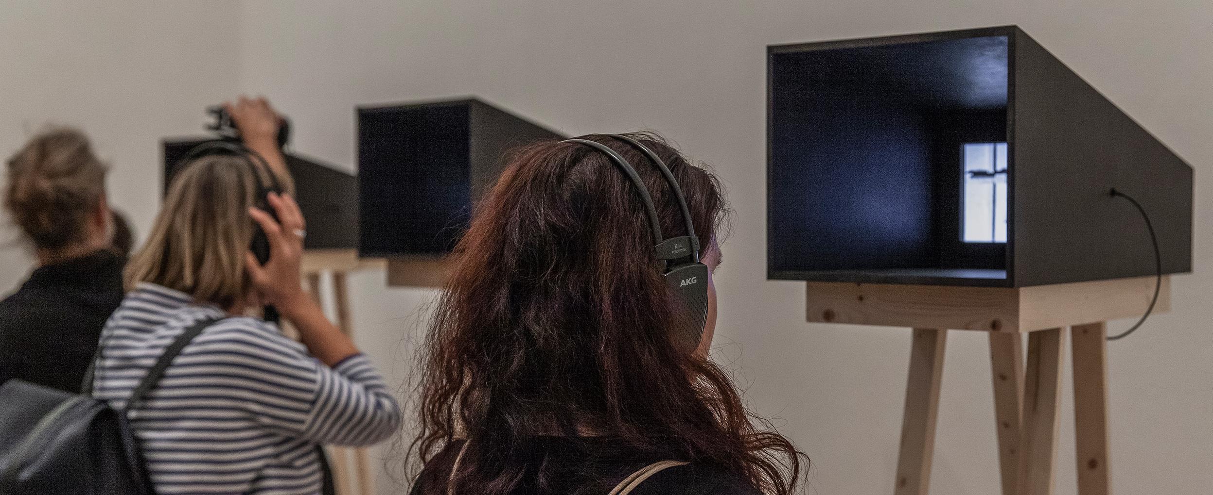 Three people each look into a video installation standing on high wooden feet.