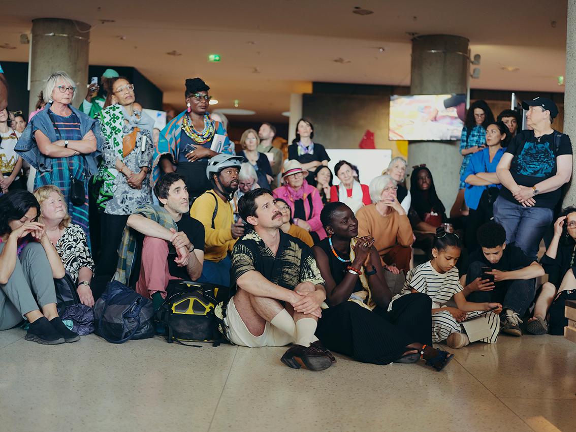 A cluster of people sits and stands in an exhibition room, looking ahead.