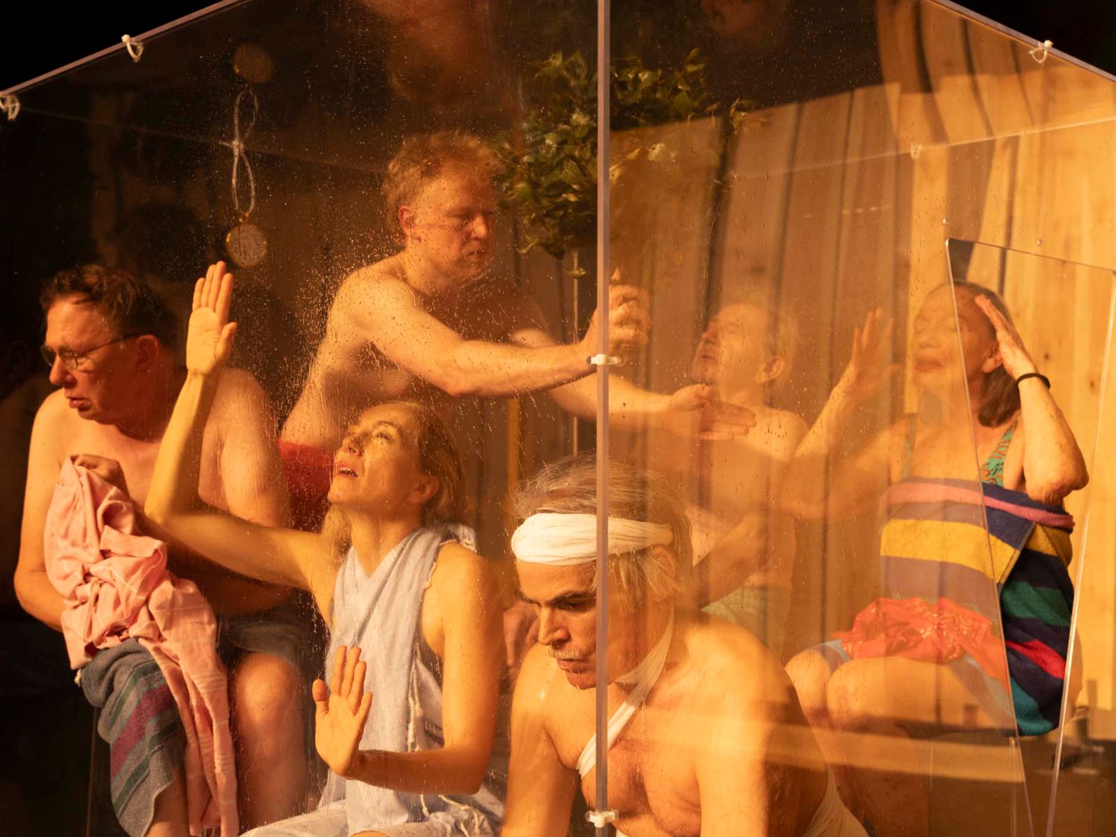 Das Ensemble sitzt in einem Glaskubus und ist in Handtücher gekleidet. Die Situation erinnert an eine Sauna.